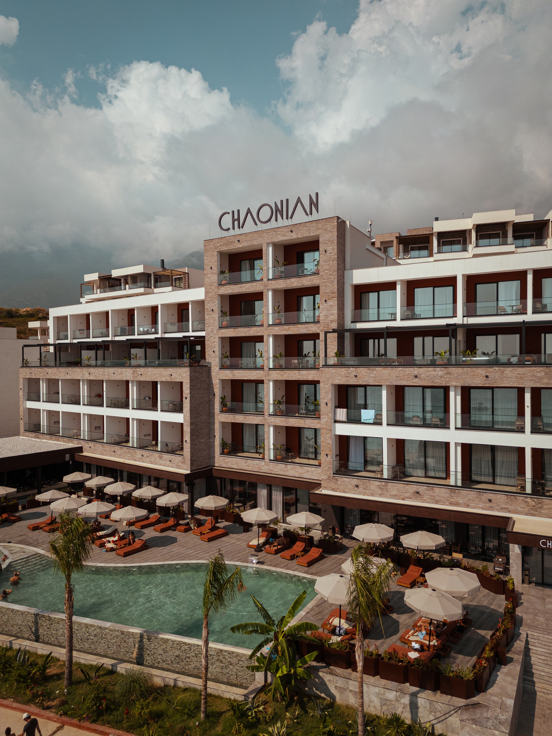 Chaonian Hotel from the air — the stone-clad facade beneath the CHAONIAN sign, the pool terrace and palms below