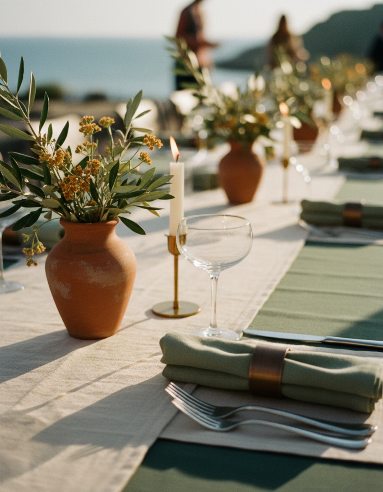 Banquet table detail — terracotta vase, olive sprigs, brass candle holder, sage napkins, sea horizon
